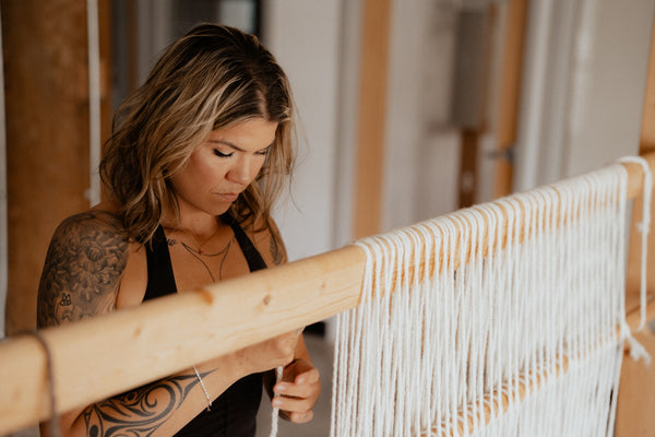 A woman with tattoos is weaving on a wooden loom in a bright, indoor studio setting.