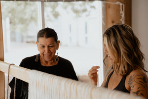 Two women collaborating in a bright, workshop, with one actively weaving on a wooden loom and the other observing closely.