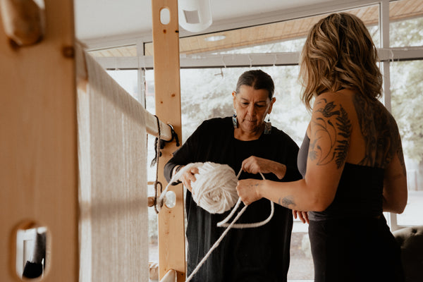 Two women working together at a large wooden loom, with one holding a ball of white yarn. The scene is set in a bright, modern studio with large windows in the background.