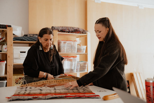 Two women in a bright, room, examining a colorful Indigenous handwoven blanket laid out on a table.