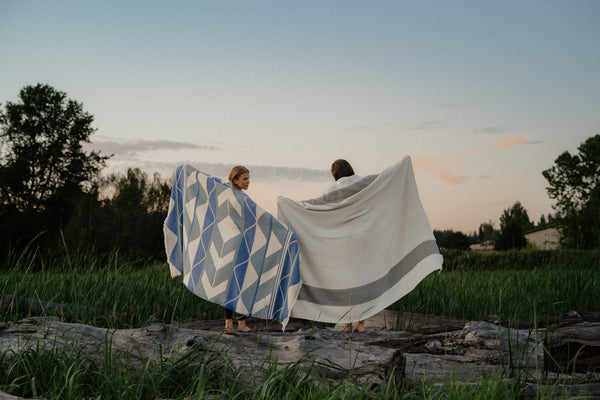 Two people standing outdoors in a grassy field at dusk, each holding up a large blanket. One blanket is white with a blue geometric pattern, and the other is white with a gray border.