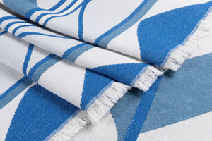 A close-up of a folded white and blue Indigenous blanket with fringed edges, showing woven texture and fabric detail.