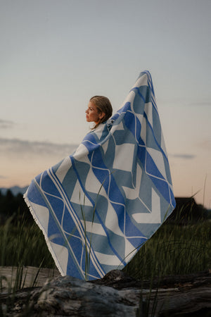 A Indigenous woman standing outdoors in a grassy field at dusk, wrapped in a large woven blanket with a blue, white, and gray pattern billowing in the wind.