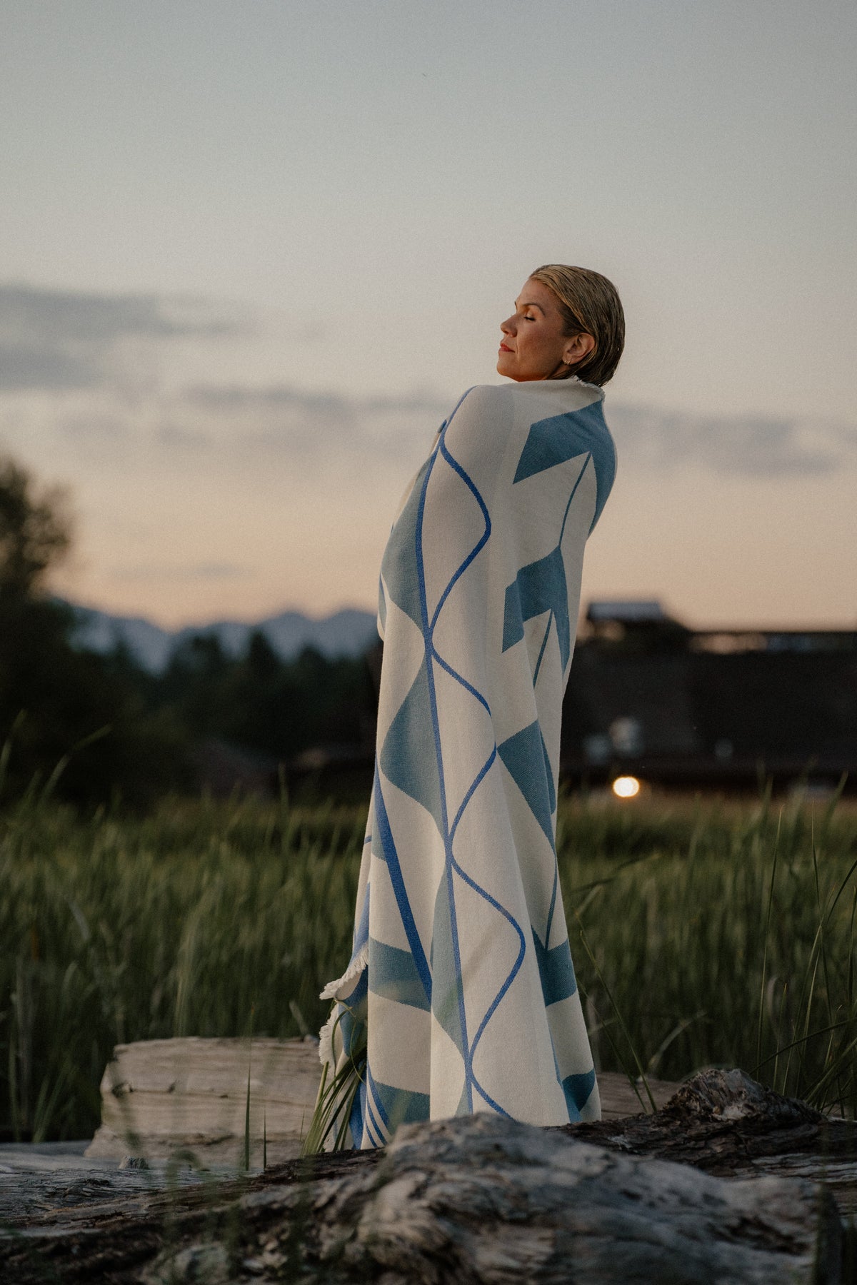 A woman stands outdoors in a grassy field at dusk, wrapped in a large white Indigenous blanket. 