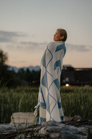 A woman stands outdoors in a grassy field at dusk, wrapped in a large white Indigenous blanket. 