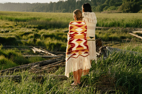 Two women standing in a grassy outdoor field, wrapped in beautiful indigenous blankets. 