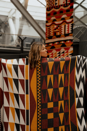A woman stands behind a large Salish woven blanket with bold geometric patterns in yellow, red, black, and white, with a matching blanket visible hanging in the background.