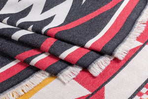 A close-up of a woven textile with fringe, featuring a bold pattern in charcoal gray, red, white, and mustard yellow. 